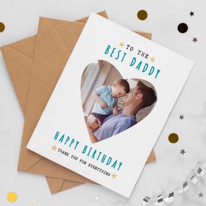 May include: A white greeting card with a heart-shaped photo of a father and child. The card reads "TO THE BEST DADDY" and "HAPPY BIRTHDAY THANK YOU FOR EVERYTHING" in teal text. The card is on top of a brown envelope.