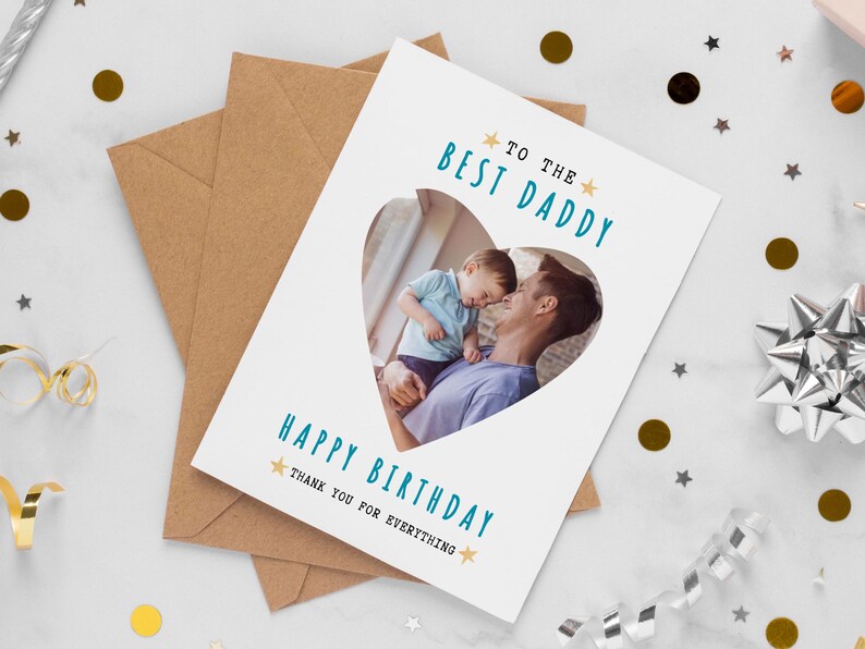 May include: A white greeting card with a heart-shaped photo of a father and child. The card reads "TO THE BEST DADDY" and "HAPPY BIRTHDAY THANK YOU FOR EVERYTHING" in teal text. The card is on top of a brown envelope.