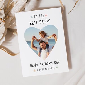 May include: A white Father's Day card with a heart-shaped photo of a father and child. The card reads "TO THE BEST DADDY" and "HAPPY FATHER'S DAY I LOVE YOU LOTS" in black text with star accents. The background is a soft, neutral color.