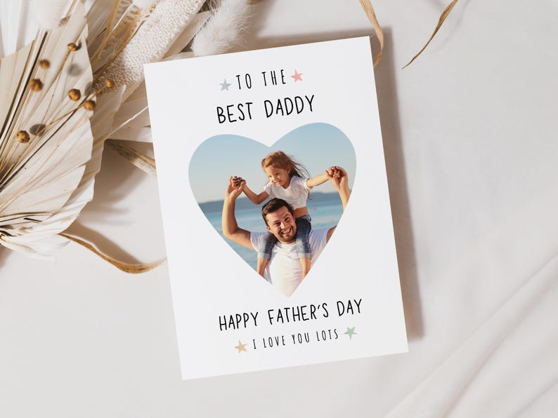 May include: A white Father's Day card with a heart-shaped photo of a father and child. The card reads "TO THE BEST DADDY" and "HAPPY FATHER'S DAY I LOVE YOU LOTS" in black text with star accents. The background is a soft, neutral color.