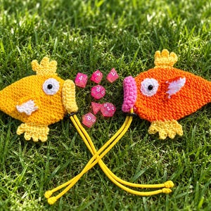 May include: Two crocheted fish pouches with yellow and orange yarn, white fins, and pink or yellow lips. Each fish features a black and white eye and a yellow drawstring cord. Between the fish are several translucent pink polyhedral dice.