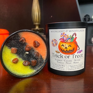 May include: A black candle jar with a Halloween-themed design featuring a pumpkin with candy spilling out. The label reads "Moonlight & Mindfulness Trick or Treat Pumpkin - Caramel - Butter Vanilla - Pecan - Maple 100% Vegan Soy Wax Crystal Candle Hand-Poured 11 oz. Therapeutic-Grade Oils Cotton-Paper Wick Crystals & Dried Flowers"