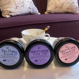 Puede incluir: Tres frascos de vidrio con tapas moradas, rosadas y azules que contienen mezclas de té de hojas sueltas. Las etiquetas son blancas con texto negro y presentan los nombres "Spa Day Calm", "Meditation Time Tea" y "Self-Care Hibiscus". Cada etiqueta incluye una descripción de los ingredientes y las instrucciones para preparar el té.