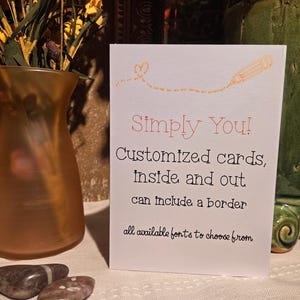 May include: A white card with the text "Simply You!" in pink and black. The card advertises customized cards with borders and font options. A pencil illustration and a heart are above the text. A vase and decorative stones are in the background.