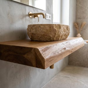 Può includere: Un lavabo da appoggio in pietra naturale con esterno ruvido e testurizzato poggia su una spessa mensola sospesa in legno chiaro con bordo naturale. Un rubinetto a parete color oro è posizionato sopra il lavabo su una parete in cemento grigio.