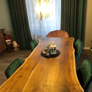 Op de afbeelding: Een grote eettafel van hout met natuurlijke randen en een warme bruine afwerking. De tafel is omringd door groene fluwelen stoelen. Een decoratief middelpunt ligt op een zwarte mat. Boven de tafel hangt een moderne kroonluchter.