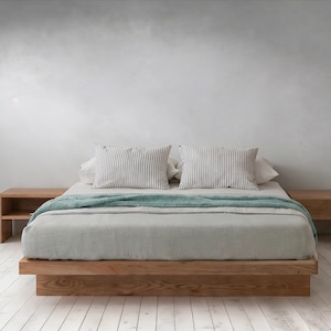Puede incluir: Una moderna cama de madera con una colcha gris claro y una manta turquesa. Dos almohadas a rayas descansan contra el cabecero. Dos mesitas de noche de madera a juego están a ambos lados de la cama. La cama está sobre un suelo de madera blanca.