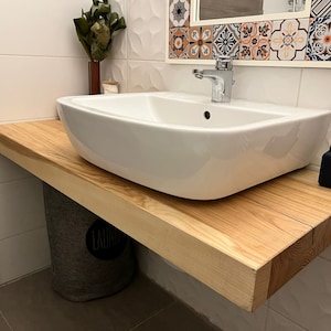 Puede incluir: Un lavabo de porcelana blanca con un grifo cromado se encuentra sobre una encimera de madera. La encimera está hecha de madera clara con veta natural. Un difusor negro con cañas se encuentra sobre la encimera a la derecha del lavabo.