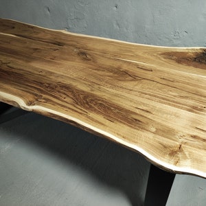 Könnte beinhalten: Ein Holztisch mit roher Kante und hellbraunem Finish. Der Tisch hat eine rechteckige Form mit einer einzigartigen, organischen Kante. Die Beine sind aus schwarzem Metall. Die Holzmaserung ist auf der Tischplatte sichtbar. Der Tisch ist ca. 180 cm lang.