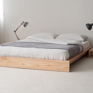 Op de afbeelding: Een lichtgekleurd houten platformbed met witte lakens en een grijze deken. Twee grijze bureaulampen flankeren het bed. Een kleine houten bijzettafel met een witte mok. Het bedframe is ongeveer 200 cm lang.