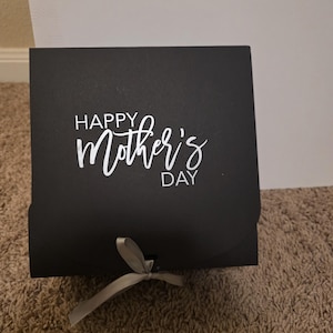 May include: A black square gift box with the words "HAPPY Mother's DAY" in white script. A silver ribbon is tied in a bow on the front of the box. The box is sitting on a brown carpet.