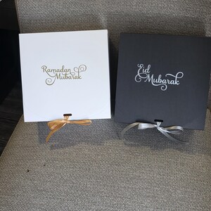 May include: Two square gift boxes, one white and one black, are displayed. The white box has "Ramadan Mubarak" in gold script, and a gold ribbon. The black box has "Eid Mubarak" in white script, and a silver ribbon. Both boxes are closed.