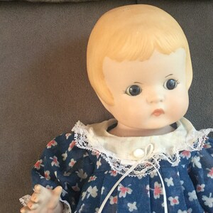 May include: A porcelain doll with blonde hair wearing a blue and white floral dress with a white lace collar.