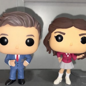 Puede incluir: Dos figuras de vinilo Funko Pop. Una figura es un hombre con un traje azul y corbata roja. La otra figura es una mujer con una chaqueta roja y una falda gris. Ambas figuras tienen ojos negros grandes y cabello castaño.