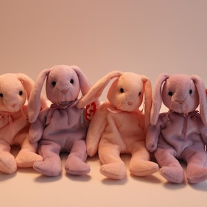 May include: Four plush pink and purple bunny beanies with Ty tags. The beanies are sitting in a row on a white surface.