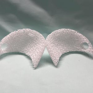 May include: Two white, semi-circular objects with a textured, woven pattern. Each has a small, circular hole near one end. The objects are displayed against a light green background.