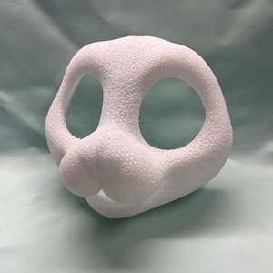 Op de afbeelding: Een witte, getextureerde konijnenmasker met grote ovale ooggaten en een afgeronde neus. Het masker is gemaakt van een solide materiaal en is ontworpen om te worden gedragen. De achtergrond is effen lichtgroen.