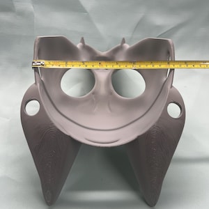 May include: A gray, 3D-printed object with a curved design, featuring two eye-like holes and a central bowl-shaped area. The object is approximately 8 inches wide, as measured by a yellow tape measure.