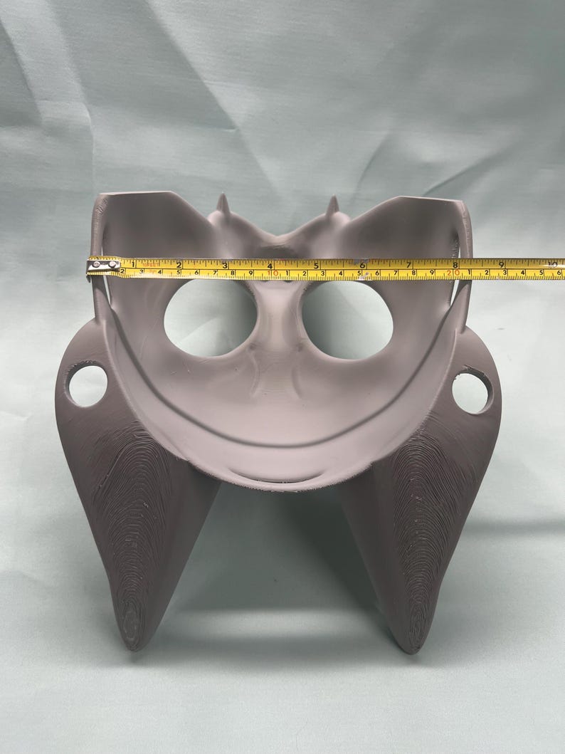 May include: A gray, 3D-printed object with a curved design, featuring two eye-like holes and a central bowl-shaped area. The object is approximately 8 inches wide, as measured by a yellow tape measure.