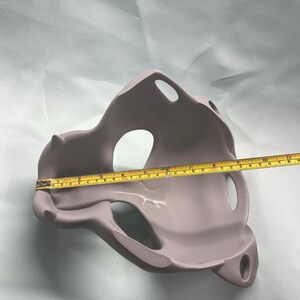 May include: A light purple, abstract-shaped object with multiple openings. A yellow measuring tape is laid across the object, showing a length of approximately 10 inches. The object has a smooth, matte finish and appears to be made of a solid material.