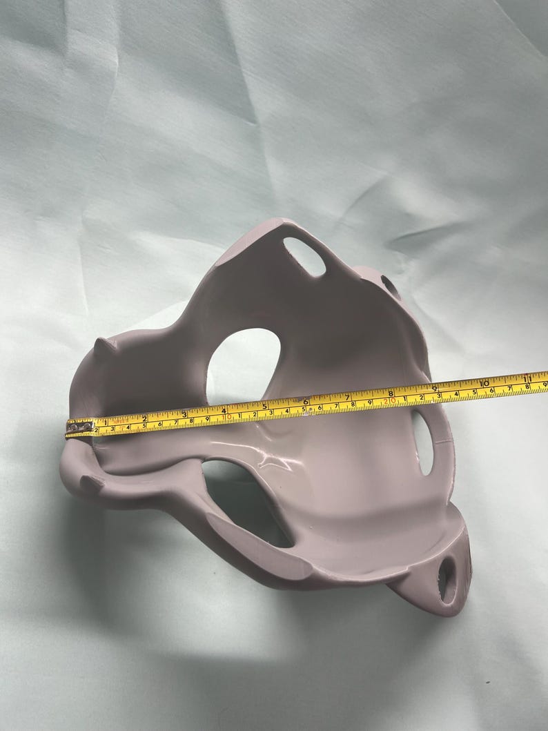 May include: A light purple, abstract-shaped object with multiple openings. A yellow measuring tape is laid across the object, showing a length of approximately 10 inches. The object has a smooth, matte finish and appears to be made of a solid material.