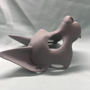 May include: A light purple cat-shaped mask with pointed ears and a prominent nose. The mask has a smooth, matte finish and is designed with eye and mouth openings. The overall shape suggests a stylized feline face, suitable for costume or artistic purposes.