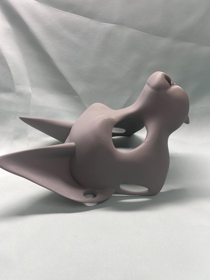 May include: A light purple cat-shaped mask with pointed ears and a prominent nose. The mask has a smooth, matte finish and is designed with eye and mouth openings. The overall shape suggests a stylized feline face, suitable for costume or artistic purposes.