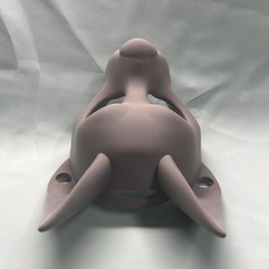 May include: A light purple, 3D-printed mask with a rounded top and two curved, horn-like projections extending from the sides. The mask has a smooth surface and appears to be designed to fit over the face, with openings for the eyes and mouth.