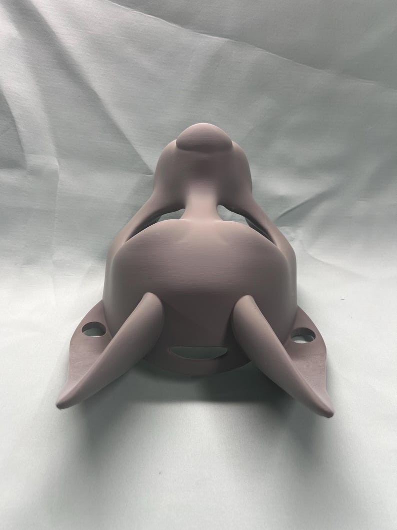 May include: A light purple, 3D-printed mask with a rounded top and two curved, horn-like projections extending from the sides. The mask has a smooth surface and appears to be designed to fit over the face, with openings for the eyes and mouth.