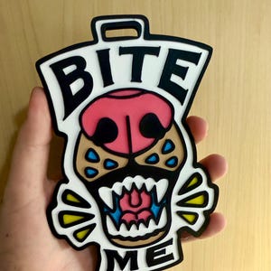 May include: A flat, multi-colored plastic tag featuring a stylized animal snout with bared teeth and a pink nose. The text BITE is printed in black at the top and ME is printed in black at the bottom. The design includes white, black, pink, blue, and yellow accents with a loop at the top for hanging.