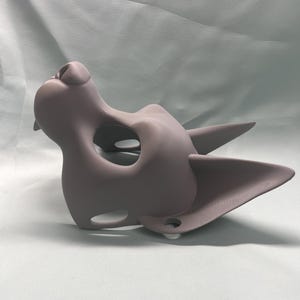 May include: A gray, cat-shaped mask with pointed ears and a prominent nose. The mask has a large eye opening and smaller holes near the mouth. The mask is made of a smooth material and is likely for costume or cosplay purposes.