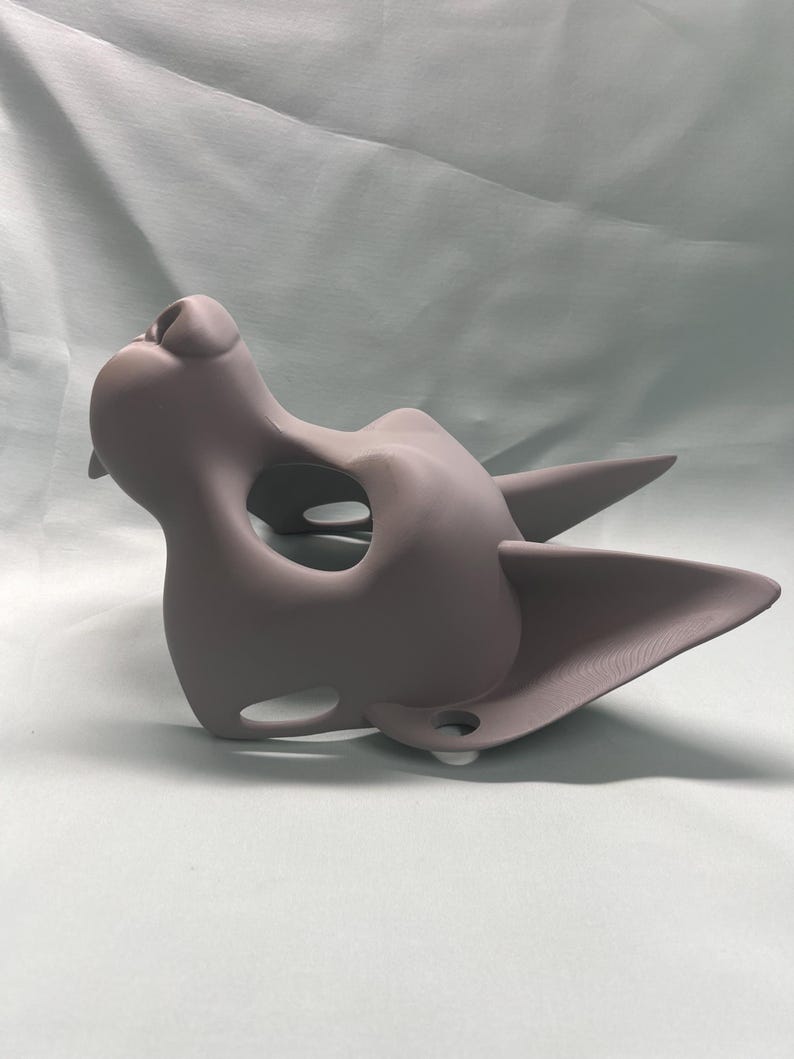 May include: A gray, cat-shaped mask with pointed ears and a prominent nose. The mask has a large eye opening and smaller holes near the mouth. The mask is made of a smooth material and is likely for costume or cosplay purposes.