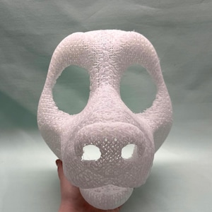 Könnte beinhalten: Eine weiße, strukturierte Maske mit großen Augenöffnungen und einer schnauzenartigen Nase. Die Maske scheint 3D-gedruckt zu sein und wird von einer Hand gehalten. Das Design der Maske ähnelt einem stilisierten Tiergesicht.