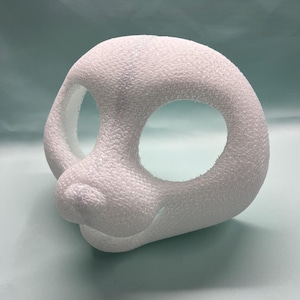 May include: A white, textured mask with large eye openings and a small nose. The mask has a rounded shape and a slight smile, suggesting a cartoon or character design. The mask is made of a material with a bumpy texture.