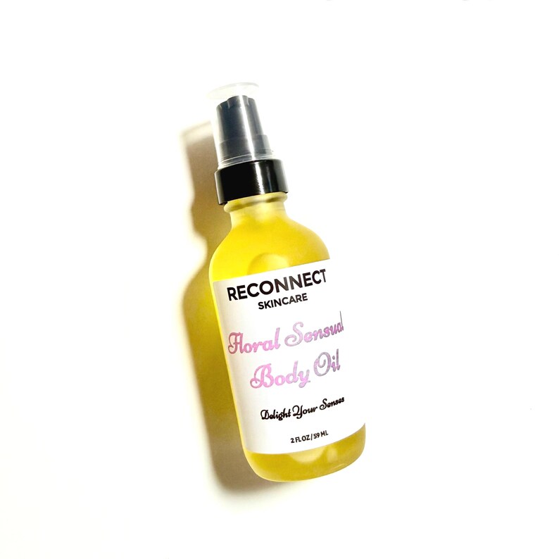 May include: A clear glass bottle filled with yellow body oil, featuring a black pump dispenser. The white label reads RECONNECT SKINCARE, Floral Sensual Body Oil, Delight Your Senses, and 2 FL OZ / 59 ML in black and pink text.