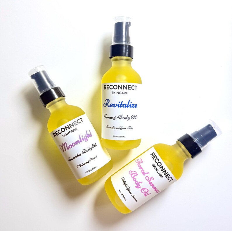 May include: Three glass bottles of Reconnect Skincare body oil with black pump dispensers. Each bottle contains yellow oil and a white label with black text, including 2 fl oz / 59 ml. Labels read Moonlight Lavender Body Oil, Revitalize Toning Body Oil, and Floral Sensual Body Oil.