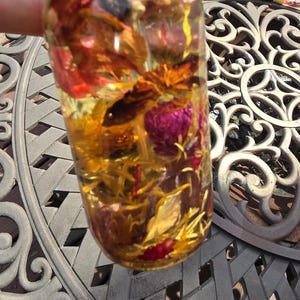 May include: A clear glass bottle filled with a golden liquid and dried flowers. The bottle contains red, yellow, and purple petals. Other bottles are visible in the background, resting on a decorative metal table.