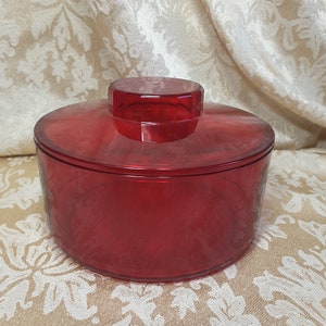 May include: A transparent red container with a matching lid. The container is round with a slightly textured surface. The lid has a geometric design and sits atop the container. The container is likely made of plastic or glass.