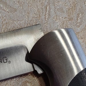 May include: Close-up of a partially visible knife with a brushed metal blade and handle. The handle has a black, textured grip. The blade has the word "ING" and a registered trademark symbol.