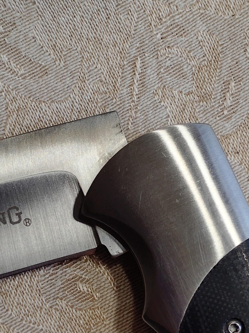 May include: Close-up of a partially visible knife with a brushed metal blade and handle. The handle has a black, textured grip. The blade has the word "ING" and a registered trademark symbol.
