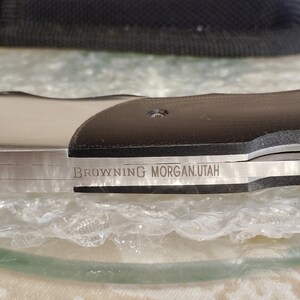 May include: A close-up shot of a knife featuring a black handle and a silver blade. The blade is inscribed with the text "BROWNING MORGAN.UTAH". The knife is displayed on a clear surface, with a black case visible in the background.