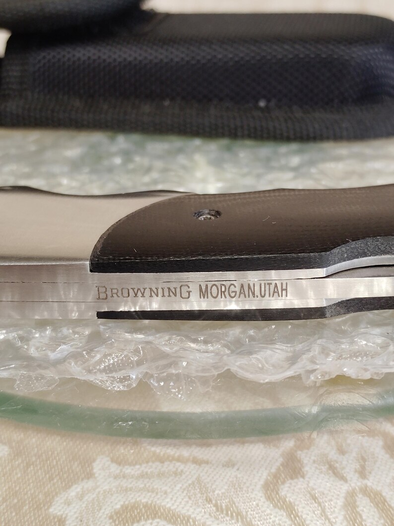 May include: A close-up shot of a knife featuring a black handle and a silver blade. The blade is inscribed with the text "BROWNING MORGAN.UTAH". The knife is displayed on a clear surface, with a black case visible in the background.