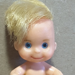 May include: A small plastic doll with a light skin tone, blue eyes, and short, blonde hair. The doll has a smiling expression and is nude. The doll's arms are slightly bent at the elbows, and its hands are in a fist.