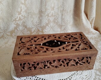 May include: A rectangular brown wooden tissue box cover featuring intricate hand-carved floral and leaf patterns across the top and sides, with a central oval opening for dispensing tissues and a decorative scalloped border along the bottom edge.