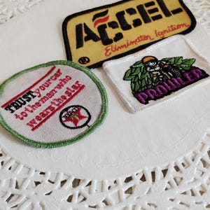 May include: Three embroidered patches: a yellow rectangular patch with black text reading ACCEL Elimination Ignition, a round white patch with green trim and red text reading TRUST your car to the man who wears the star, and a white rectangular patch with a graphic of a figure and the word PROWLER in purple.