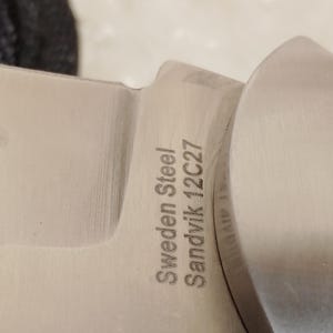 May include: Close-up of a stainless steel knife blade. The text "Sweden Steel Sandvik 12C27" is etched into the metal. The blade has a curved edge and a polished finish.