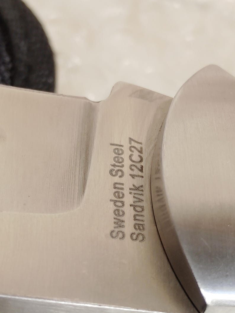 May include: Close-up of a stainless steel knife blade. The text "Sweden Steel Sandvik 12C27" is etched into the metal. The blade has a curved edge and a polished finish.
