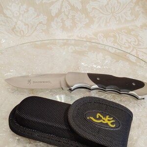 May include: A stainless steel folding knife with a black handle and the word "Browning" printed on the blade. A black nylon sheath with a yellow logo is also included.