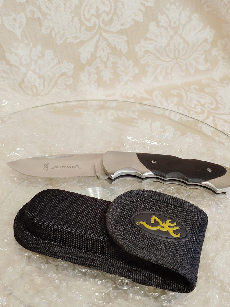 May include: A stainless steel folding knife with a black handle and the word "Browning" printed on the blade. A black nylon sheath with a yellow logo is also included.