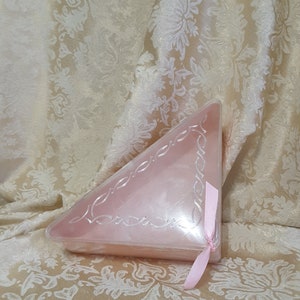May include: A pink, triangular-shaped box with a decorative silver border and a pink ribbon. The box is sitting on a cream-colored fabric with a floral pattern. The box appears to be made of plastic.